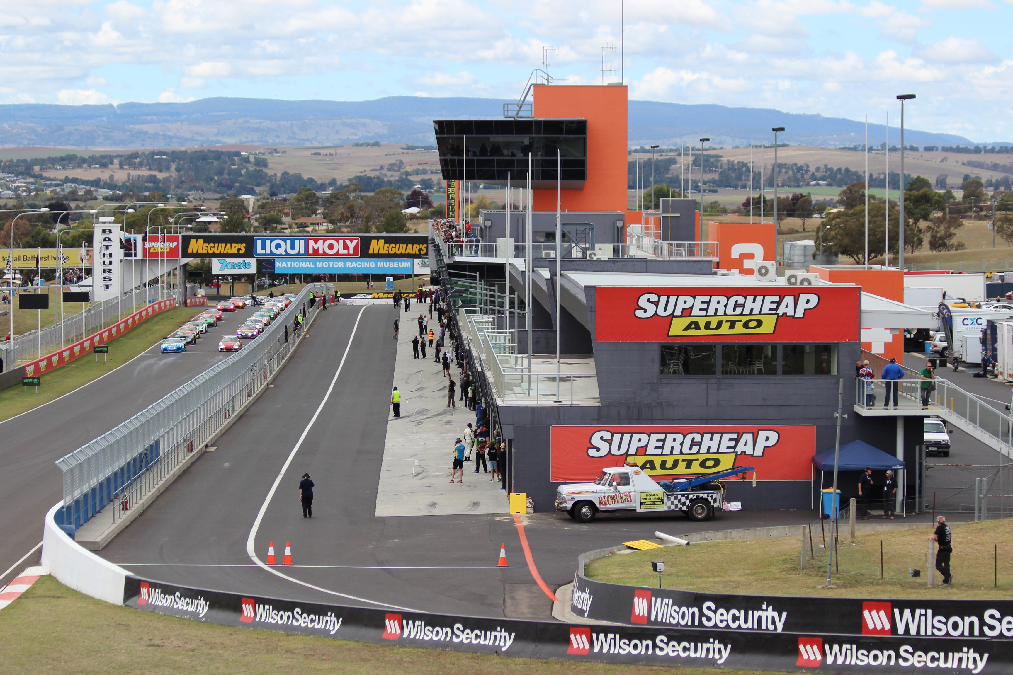 levated view of the pit straight and pit lane at Mount Panorama, Bathurst, in 2015. A line of racing cars approaches the start-finish area, with the modern pit complex, garages and distinctive orange race control tower dominating the foreground. The surrounding countryside and distant hills provide a backdrop to the circuit.